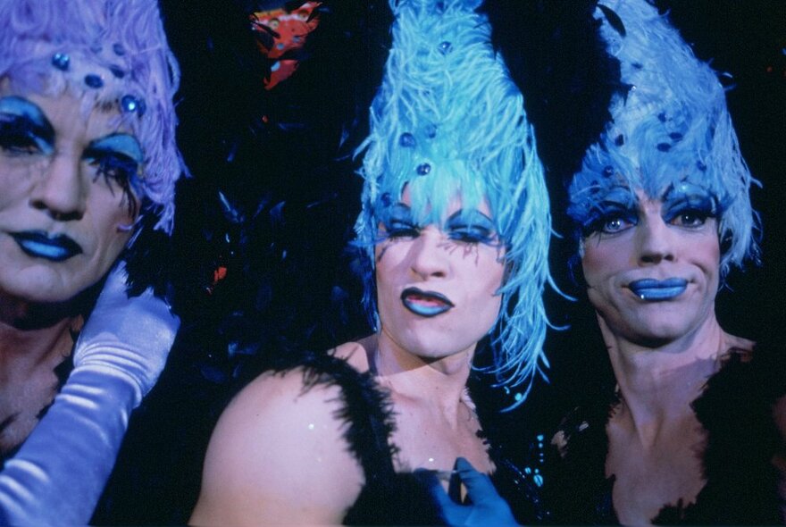 Close-up of the three actors from the film, The Adventures of Priscilla, Queen of the Desert, all wearing drag make-up, feathered wigs and satin gloves.