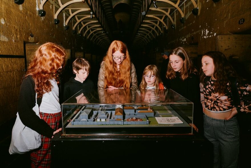 A group of children surrounding an exhibit in a glass case beneath the cell walkways at Old Melbourne Gaol.