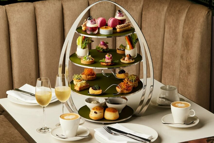 Four-tiered, egg-shaped platter featuring petits-four and small desserts; two flutes of champagne and one cup of coffee to left, cup of coffee to right; brown banquette behind.