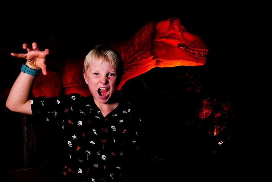 A young boy holding up his hand like a claw, with mouth open, in front of a lit up dinosaur at Melbourne Zoo after dark.