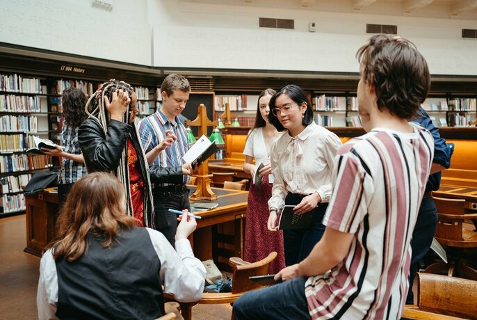 A group of young adults in an activity that involves looking at or reading from books in a library setting.