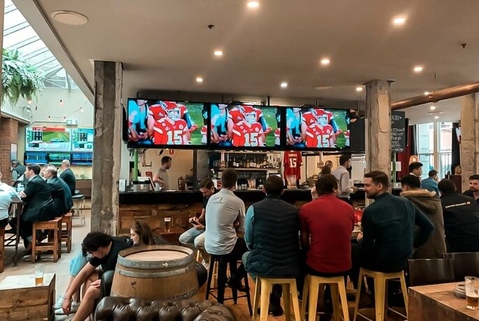 Interior of King Hotel showing the bar area with  people seated on high stools, and three large televisions screening live sport.