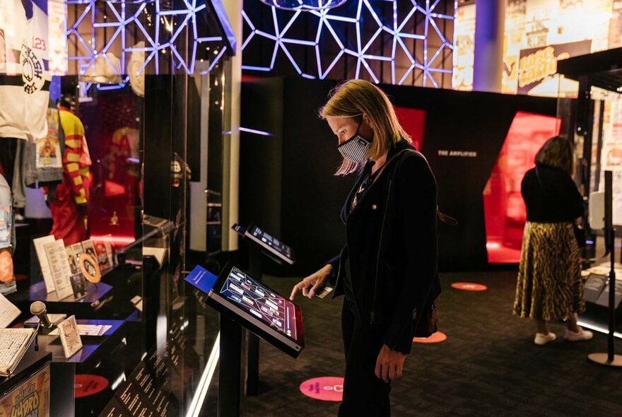 A museum visitor in an exhibition space using a screen, surrounded by display objects.