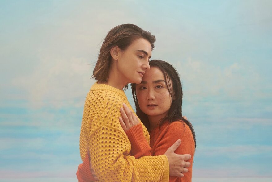 Two people embracing, dressed in yellow and orange with the ocean in the background.