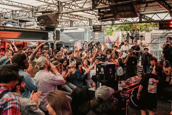 Crowd of onlookers enjoying themselves and applauding in an outdoor club or bar, watching a person rapping into a microphone next to a DJ mixing desk of turntables.