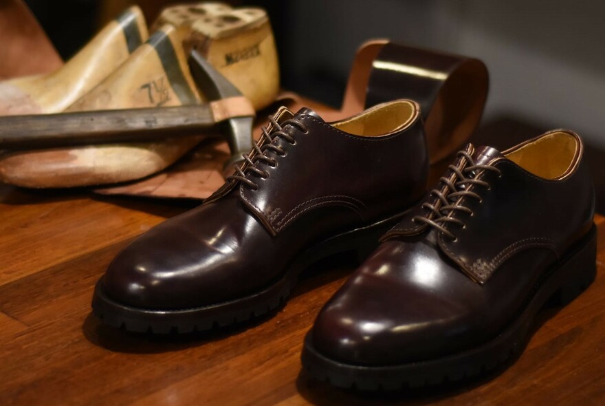 A pair of shiny lace-up brown leather shoes on a wooden table with wooden shoe lasts in the background.