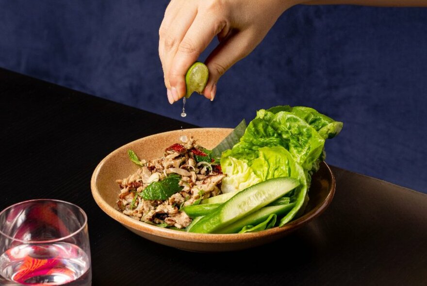 Hand squeezing fresh lime onto a bowl of food, with a salad on the side of the bowl.