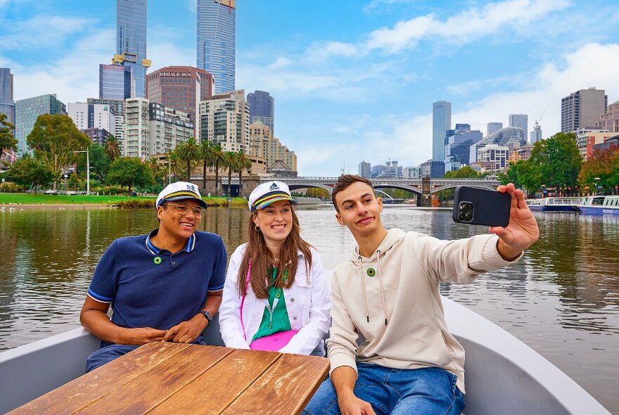 Three people sitting at the back of a small boat taking a selfie, while travelling down a city river.