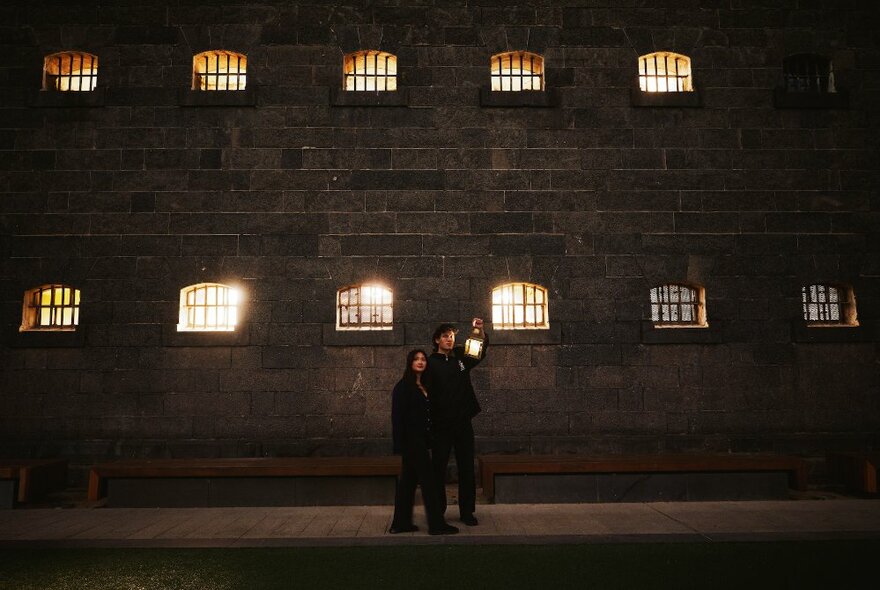 Two people outside Old Melbourne Gaol at night, the lights inside visible through tiny windows.