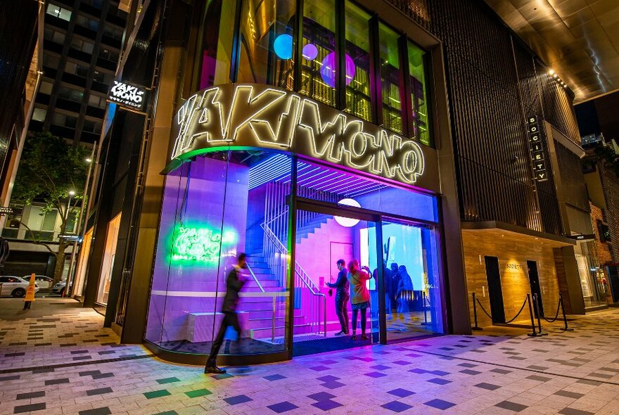 People entering a restaurant with neon lights and a sign reading 'Yakimono'.