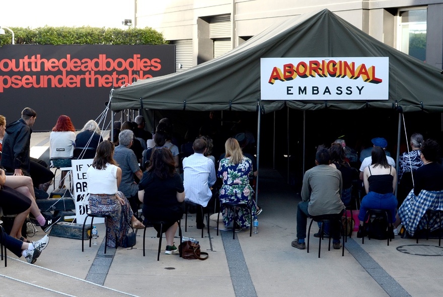 People seated in chairs in an open space, in front of a marquee that has a large sign on the front of it that reads ABORIGINAL EMBASSY.