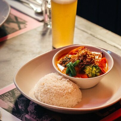 A white bowl with a neat mound of white rice, and a smaller bowl of Thai massaman curry, with a glass of beer in the backgound.
