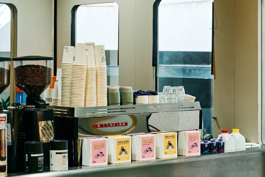 Service counter of a small cafe space showing a coffee making machine, paper takeaway cups and packets of tea or coffee for sale.