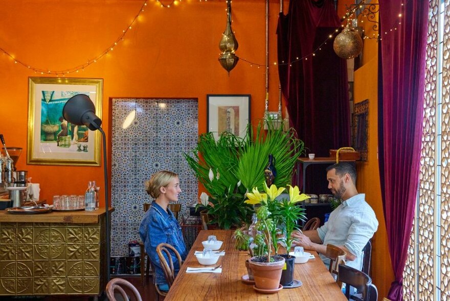 Restaurant interior showing a couple dining at a long wooden table, an orange wall behind them, Moroccan tiles, plants and decorations surrounding them.