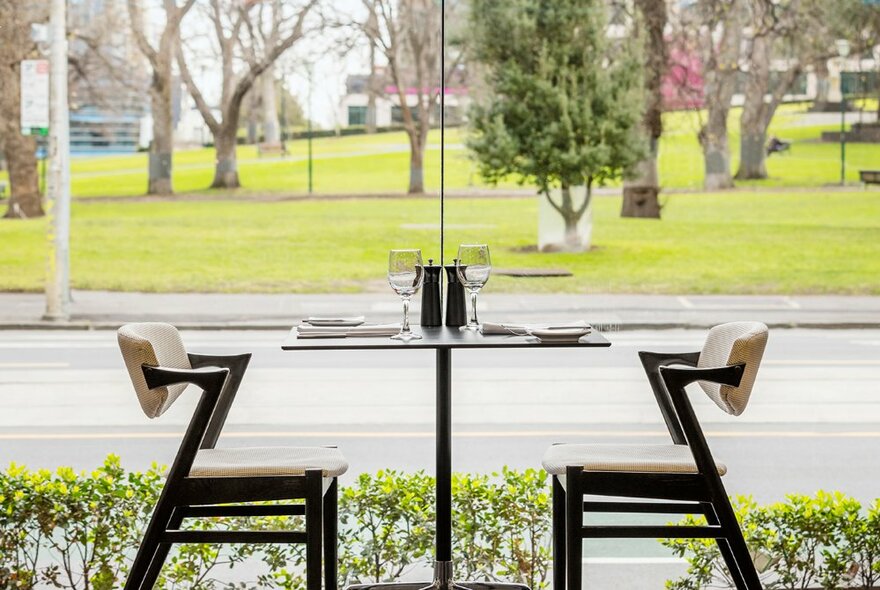 Restaurant table and chairs positioned next to full-length windows overlooking a road and parkland.