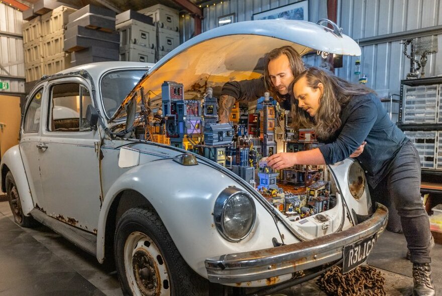 Old blue car with engine open to reveal figurines within, two people leaning over.