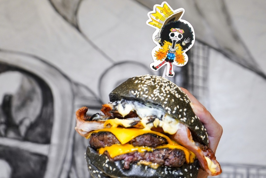 A burger being held with one hand featuring a black seeded bun, two slices of cheese, two meat patties and an anime character stuck in the top. 