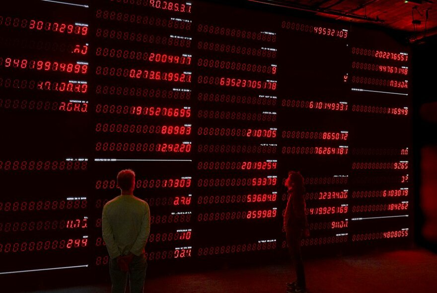 People standing in front of a large screen installation displaying lines of red numbers. 