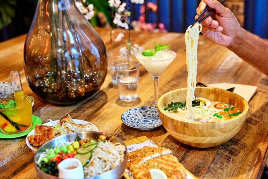 A hand lifting noodles out of a bowl using chopsticks, with other plates of food and drinks on the dining table. 