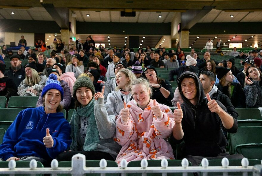 People in warm clothes and oodies at the MCG with their thumbs up. 
