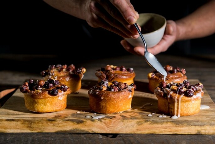 Six small pastries topped with fruit, hands above drizzling icing with a spoon.