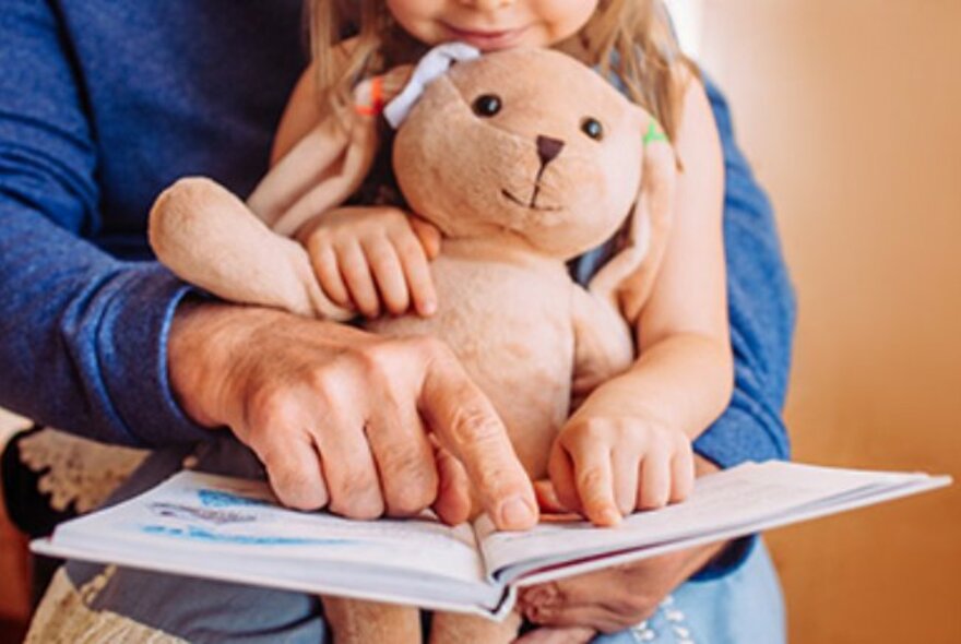 A child with teddy bear seated on an adult's lap, their fingers pointing at a page in a picture book.