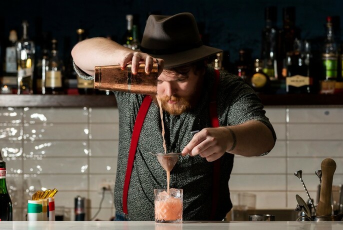 Bartender behind the bar pouring a mixed drink from a cocktail shaker into a glass.