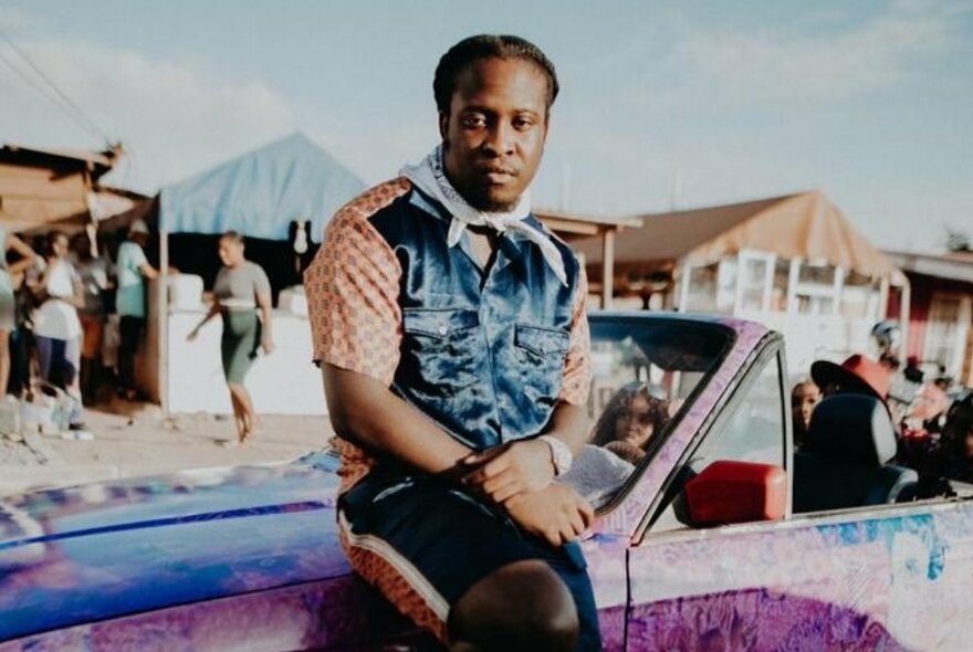 English musician, Kojo Funds, wearing a colourful outfit leaning against a colourful convertible car at a music festival-type event.
