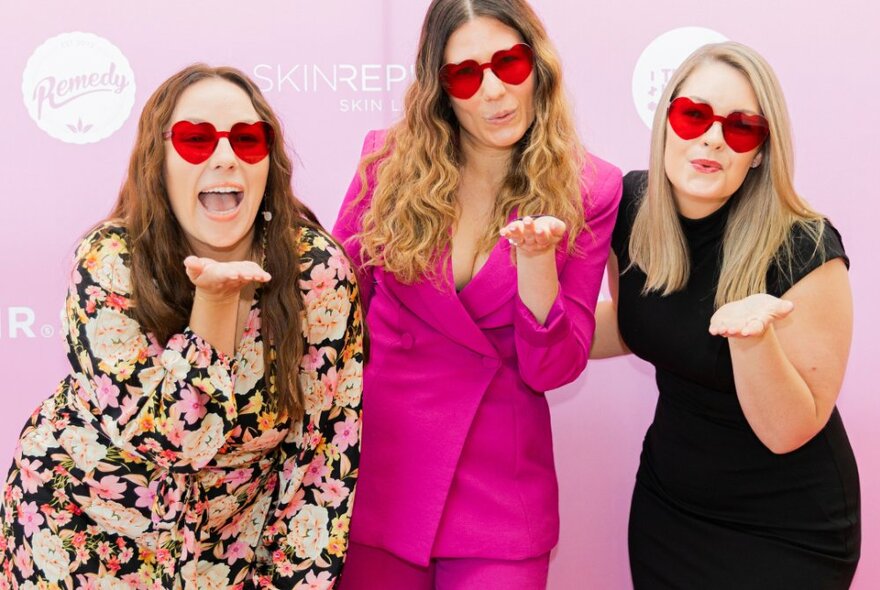 Three women indoors wearing red heart shaped sunglasses all in the process of blowing a kiss.
