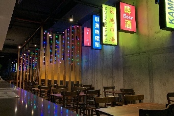 Interior of restaurant showing tables and chairs against a concrete wall, and illuminated signs hanging from the wall.