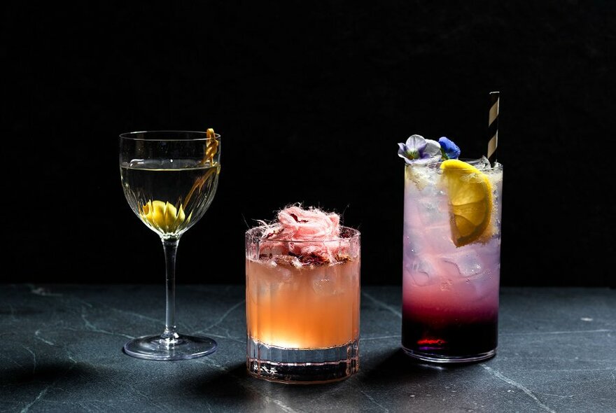 Three colourful backlit cocktails on a dark tabletop.