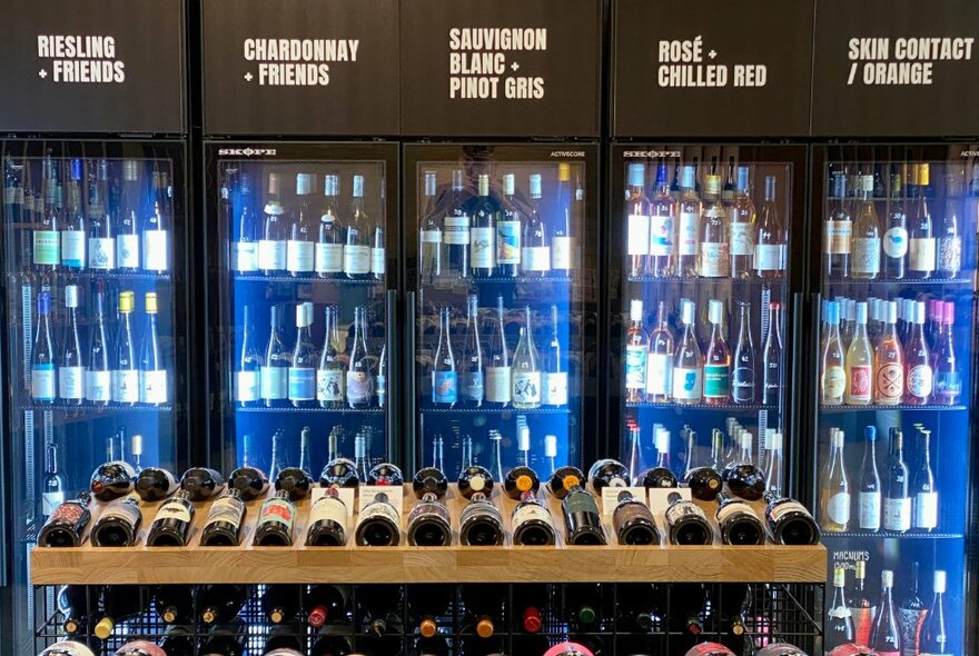 A wooden wine shelf with wine bottles displayed, in front of a five-section wine fridge with tens of bottles of wine.