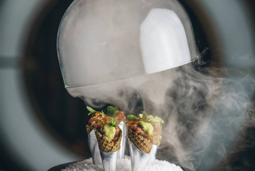 Glass lid being lifted off to reveal platter of waffle cones amid dry ice mist.