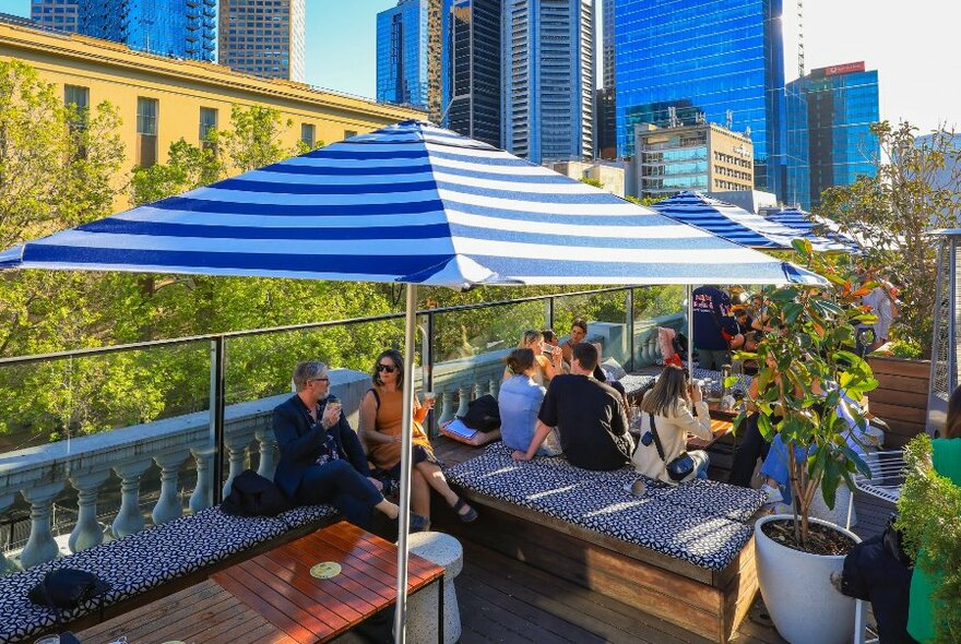 People enjoying the shade under blue and white umbrellas on a rooftop bar.