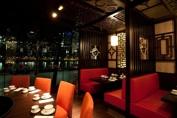 Moody restaurant interior with downlit round table, red banquettes in booths with dark wood screens beside night view of Yarra River.