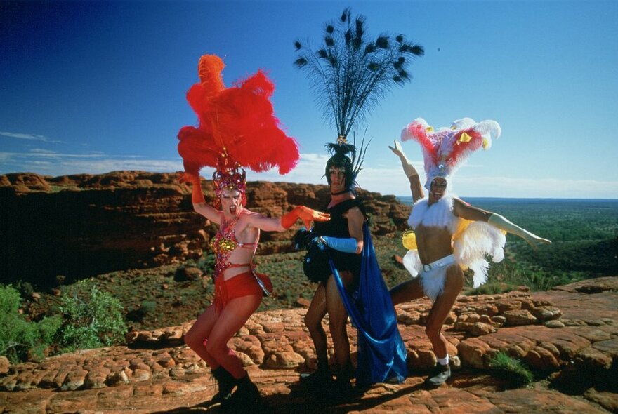 The three characters from the film The Adventures of Priscilla, Queen of the Desert, in drag costumes, standing on a flat red cliff top in the Northern Territory outback.