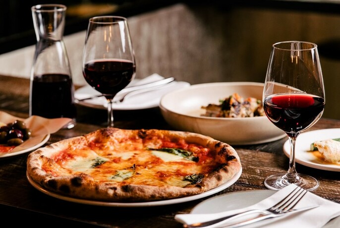 A table set for two with a pizza on one plate and a pasta dish on the other, with two glasses and a carafe of red wine.