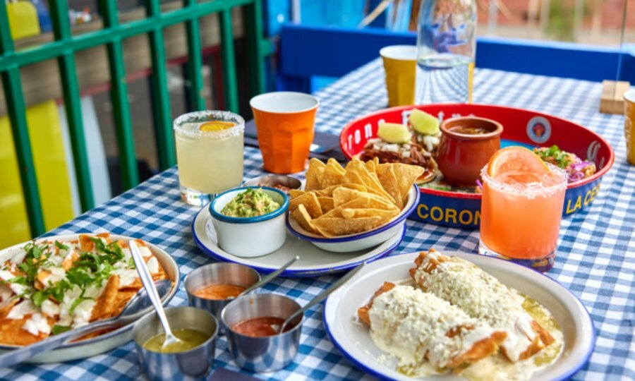 Full table of food, nachos, corn chips with guac, enchiladas, condiments and cocktails. 