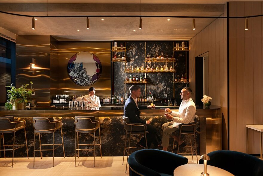 The softly lit bar at the Dorsett Melbourne with two people seated on high bar stools having a drink at the counter, and a bartender behind the bar.