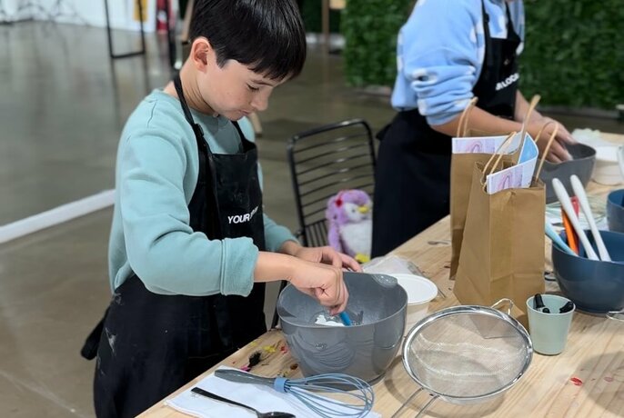 Two children wearing aprons mixing ingredients in a bowl on a workbench, and participating in a cooking workshop.