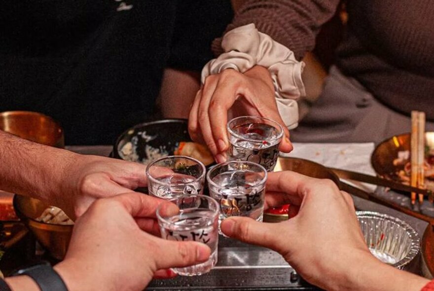 Four hands holding four small shot glasses filled with a clear spirit, clinking the glasses together over a table of food.