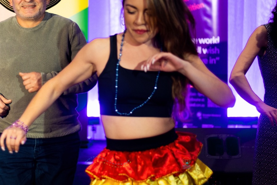 Woman seen from forehead to just below waist, with long, dark hair, wearing black tank top, blue beads and red and yellow ruffled skirt, arms slightly raised, dancing.