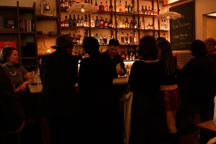 Moodily lit dark bar with customers seated in front of the backlit bar and bartender.
