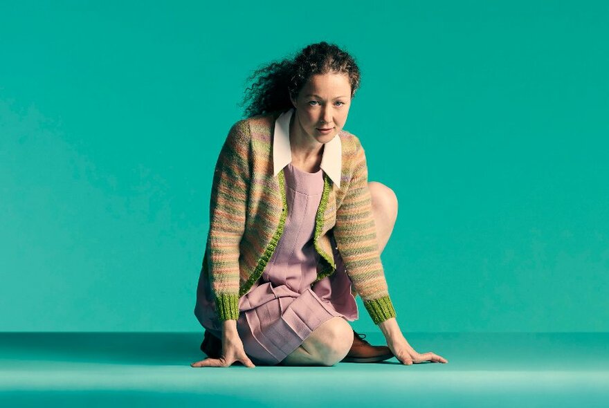 Young adult with long hair tied back into a ponytail and wearing clothes in the style of the late 1950s (pink pleated dress, white collar, hand-knitted cardigan), half-seated or crouched on the ground against a mint green backdrop.