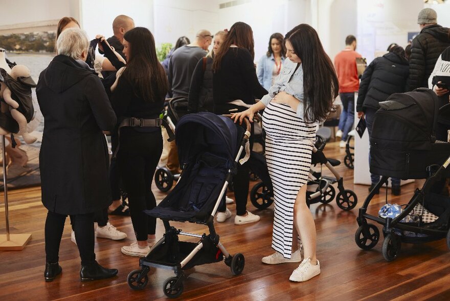 Pregnant woman looking at a pram in an event space with people in the background looking at stalls with products on display.