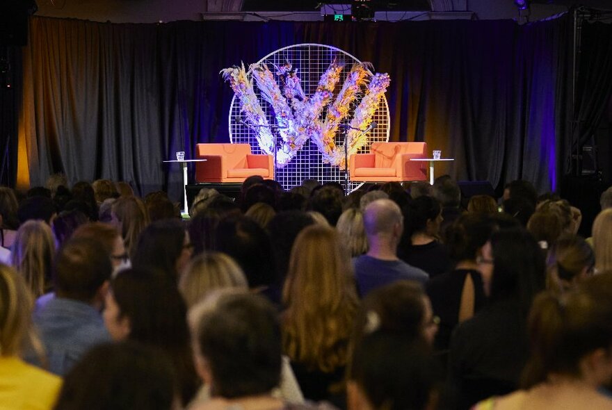A seated audience in a room, seen from behind, they all face the stage where there are two empty armchairs and a backdrop of a double W sign.
