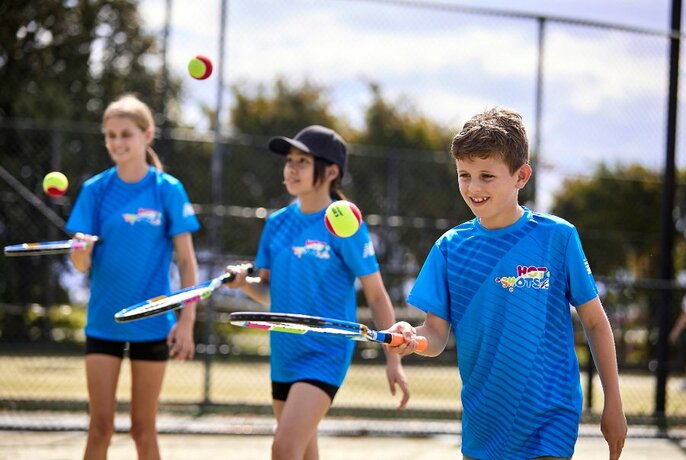 Three children in matching blue t-shirts practise hitting tennis balls upwards on their racquets. 