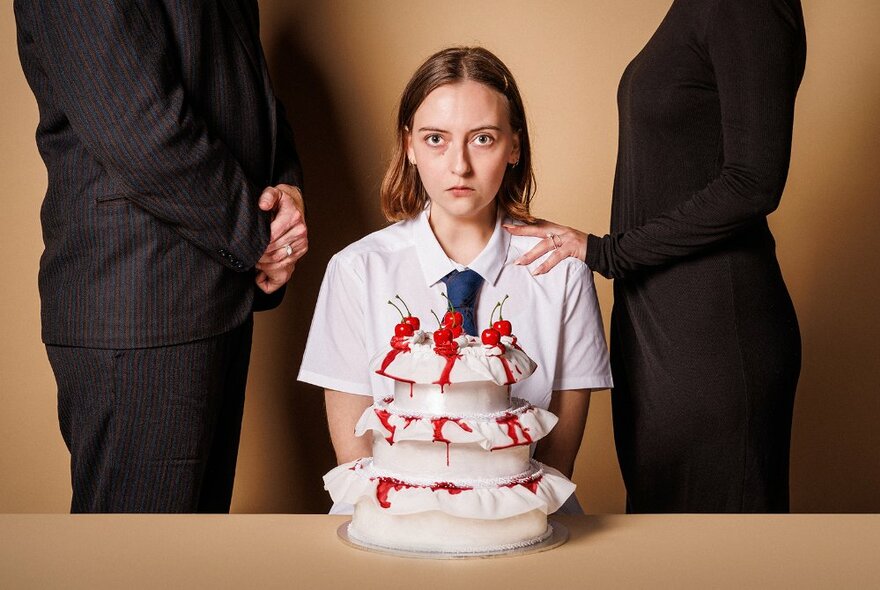 A girl seated in front of a blood-spattered cake, her parents in black standing guard.