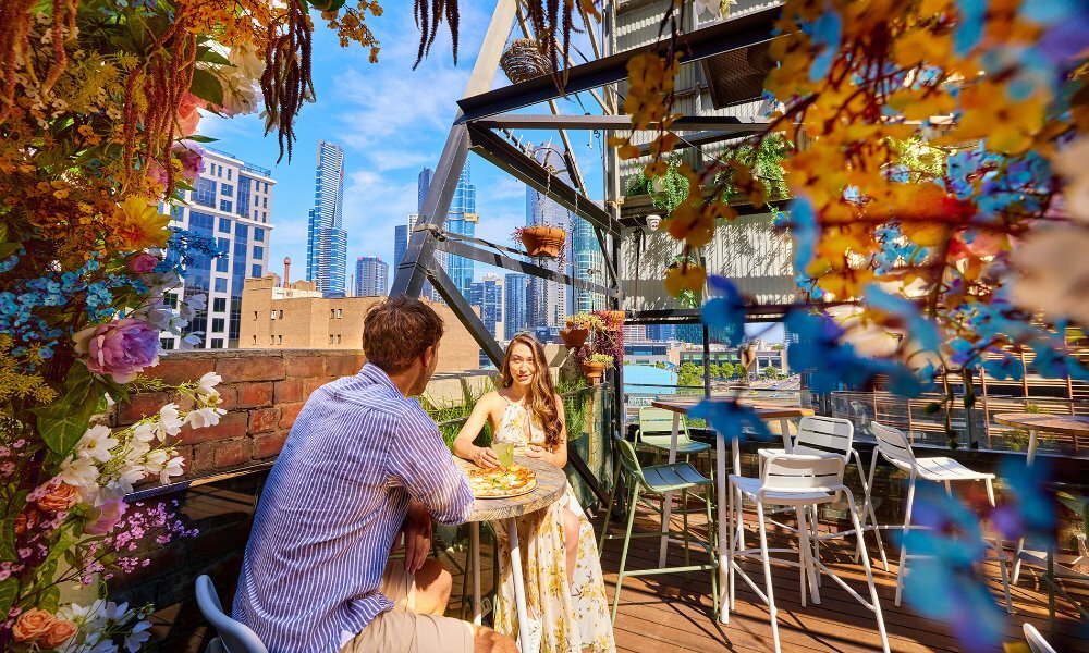 A couple sharing pizza surrounded by hanging flowers at a rooftop bar.