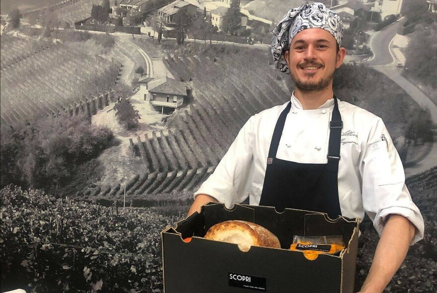 Chef holding a box of bread and produce and standing in front of a large black and white image of a vineyard.